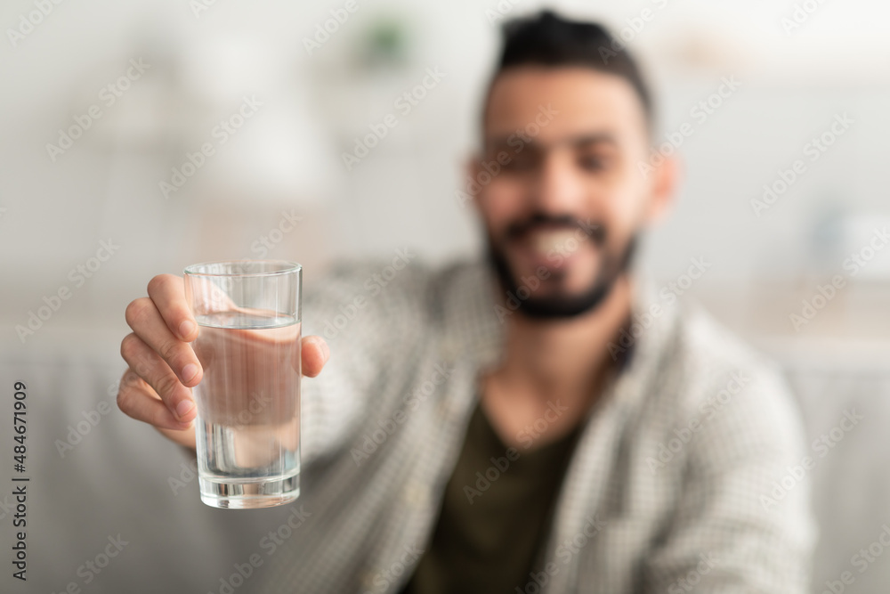 Young Arab man showing glass of fresh clear water at camera at home, selective focus. Wellness concept