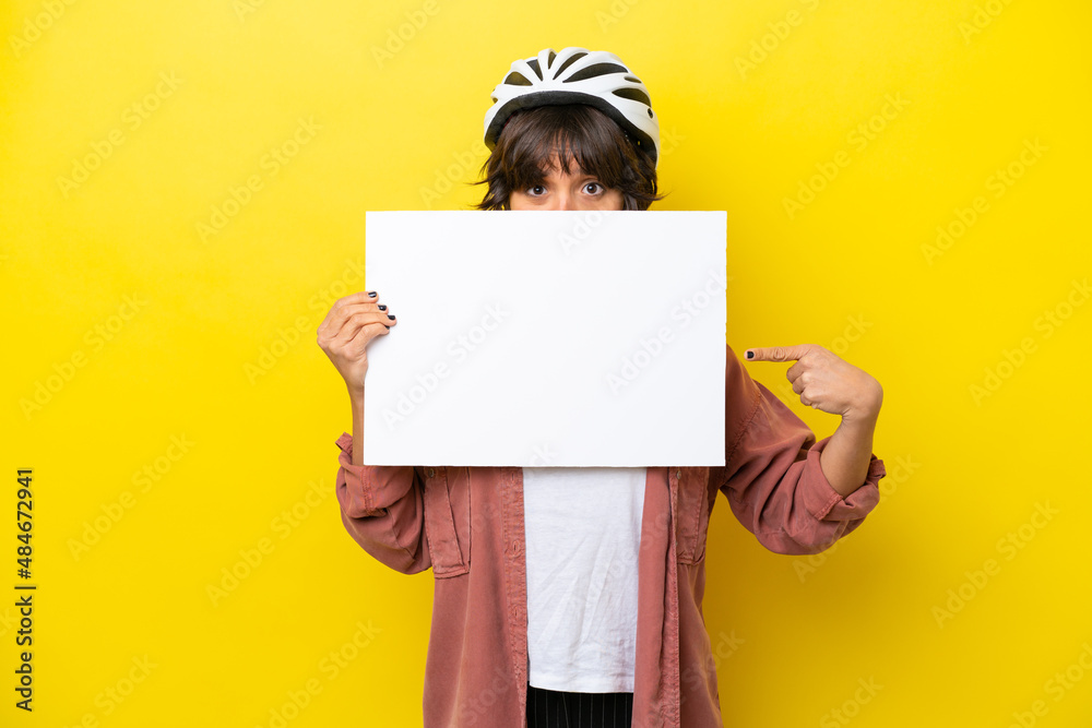 Young cyclist latin woman isolated on yellow background holding an empty placard and hiding behind it Young cyclist latin woman isolated on yellow background holding an empty placard and hiding behind it