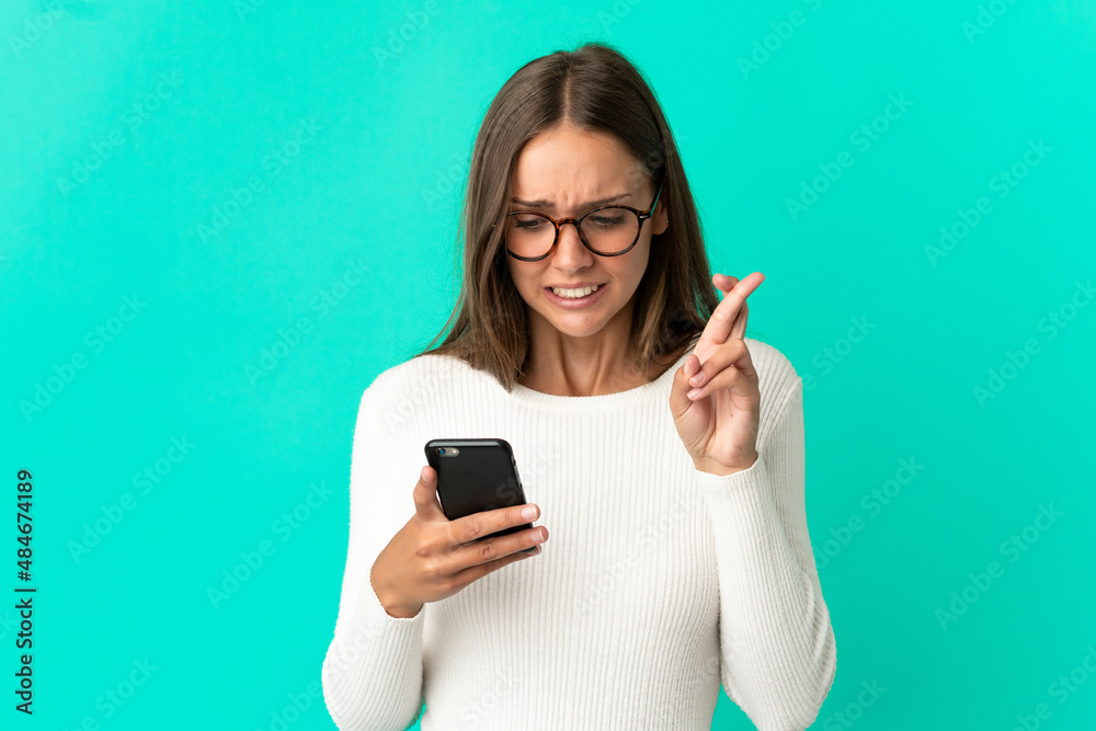 Young woman over isolated blue background using mobile phone with fingers crossing Young woman over isolated blue background using mobile phone with fingers crossing