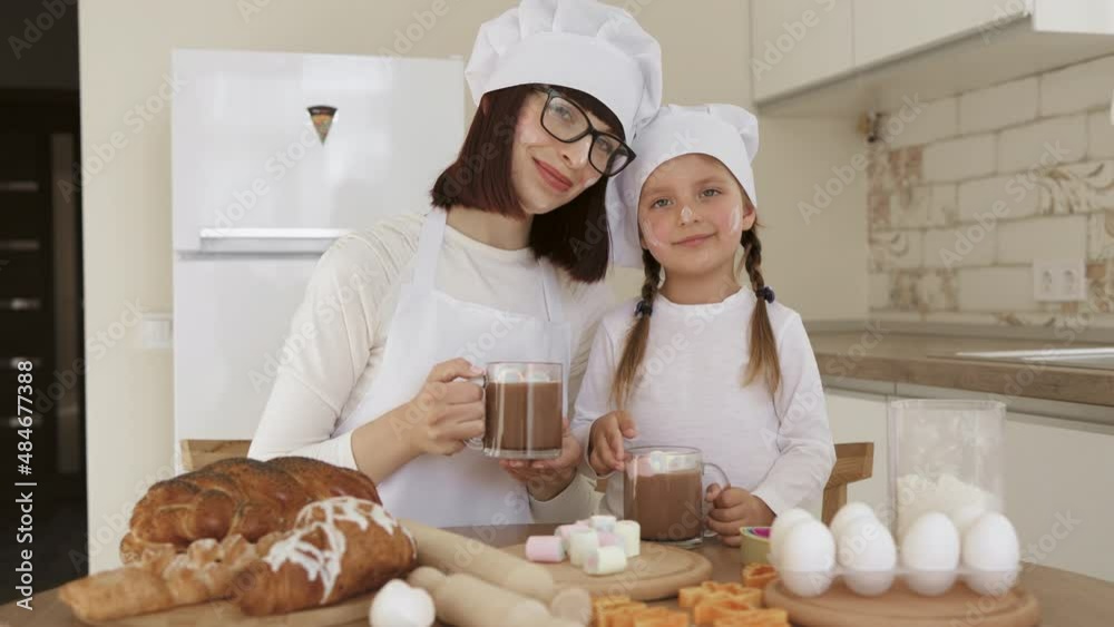 Cute little caucasian girl and her beautiful mom in white hats of chef and aprons and faces smeared with flour drinking hot cocoa with marshmellow and looking at camera at home Cute little caucasian girl and her beautiful mom in white hats of chef and aprons and faces smeared with flour drinking hot cocoa with marshmellow and looking at camera at home