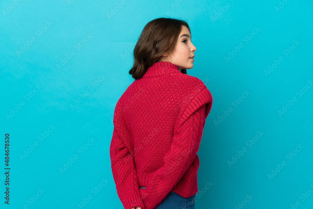 Little caucasian girl isolated on blue background in back position and looking back Little caucasian girl isolated on blue background in back position and looking back