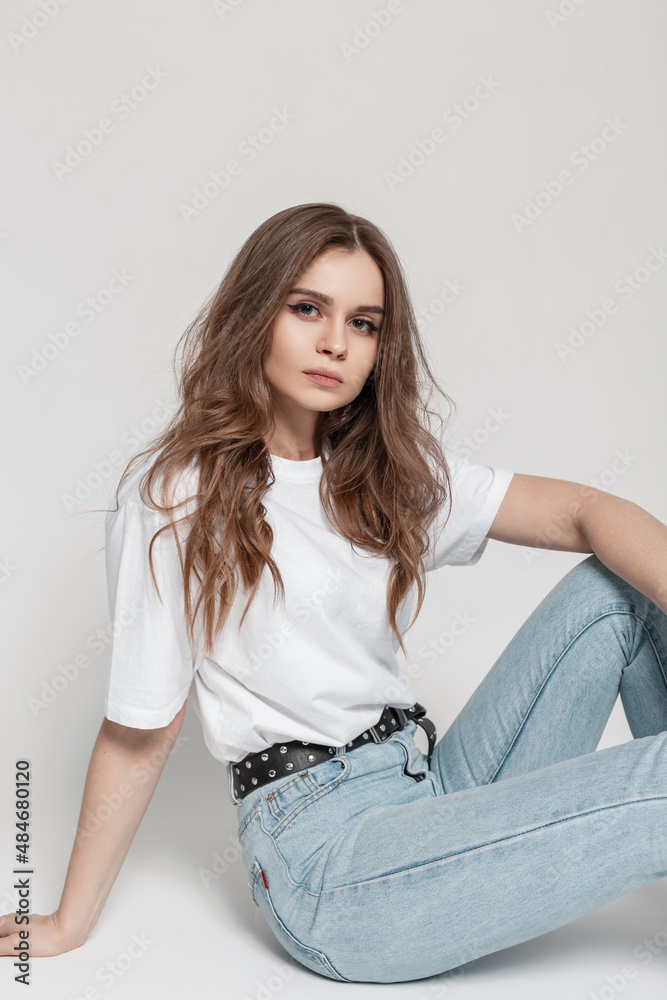 Stylish beautiful young girl with a hairstyle in fashionable clothes with a  white T-shirt and blue vintage jeans sits in the studio Stock Photo | Adobe  Stock