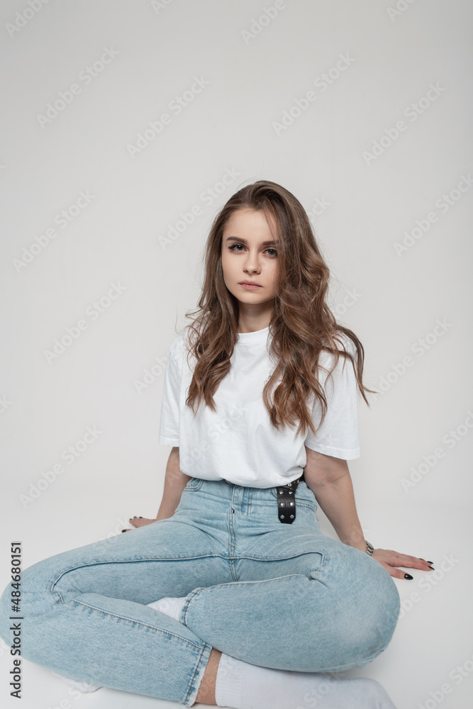Fashionable pretty young woman model in a white t-shirt and blue jeans sits in the studio on a white background Fashionable pretty young woman model in a white t-shirt and blue jeans sits in the studio on a white background