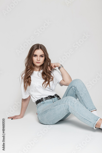 Glamorous beautiful serious girl model in casual clothes with a white t-shirt and blue high waist jeans sits in the studio