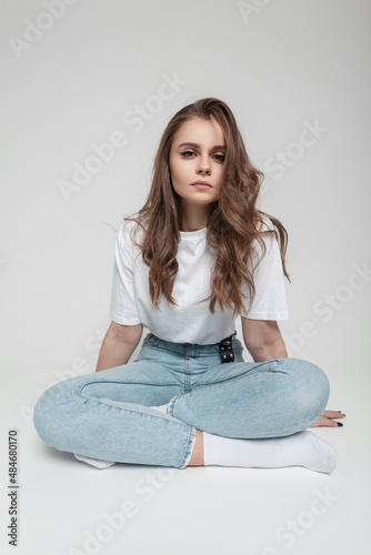Beautiful young model girl with curly hair in a trendy white mock up T-shirt with jeans and white socks sitting in the studio on a white background