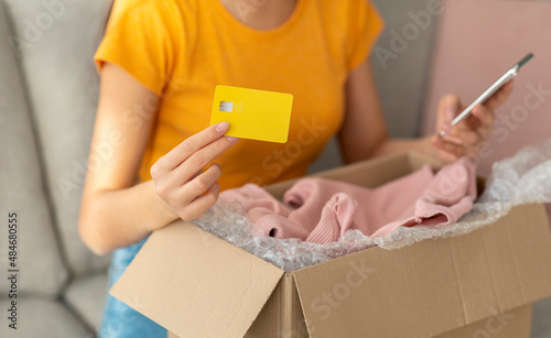 Delivery concept. Young lady holding credit card and using smartphone, shopping online from home, sitting on sofa © Prostock-studio