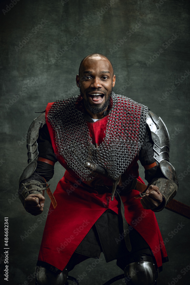 Half-length portrait of dark skinned man, medieval warrior or knight wearing armour shouting as winner isolated over dark vintage background. Comparison of eras, history