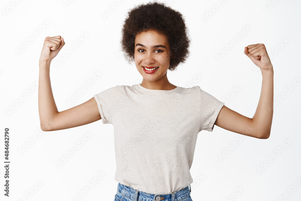 Strength and women workout concept. Smiling black girl with afro hair, young woman flexing biceps, showing muscles on arms, standing over white background