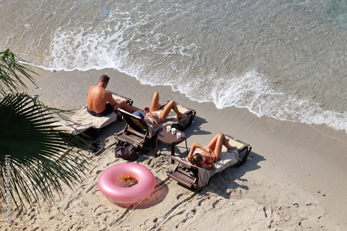 Fototapeta Naklejka Na Ścianę i Meble -  Man and two young women in bikini sunbathing on a sandy beach, top view. People with smartphones lying in deck chairs, vacation on a sea resort