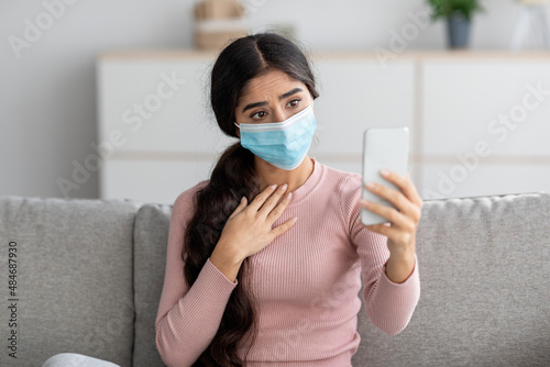 Sad young indian lady in protective mask on sofa makes selfie on smartphone or video call in living room
