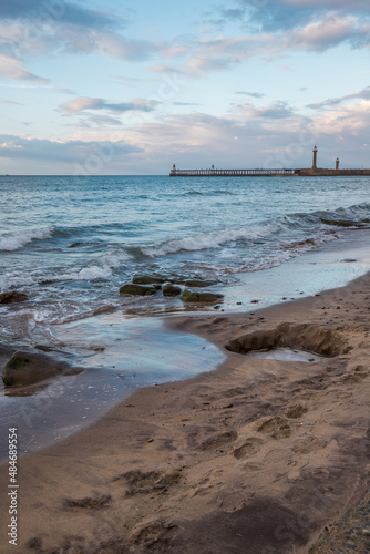 Evening light at Whitby beach. Warm sky and gentle waves along a sandy coastline with large rocks. Whitby pier in the background