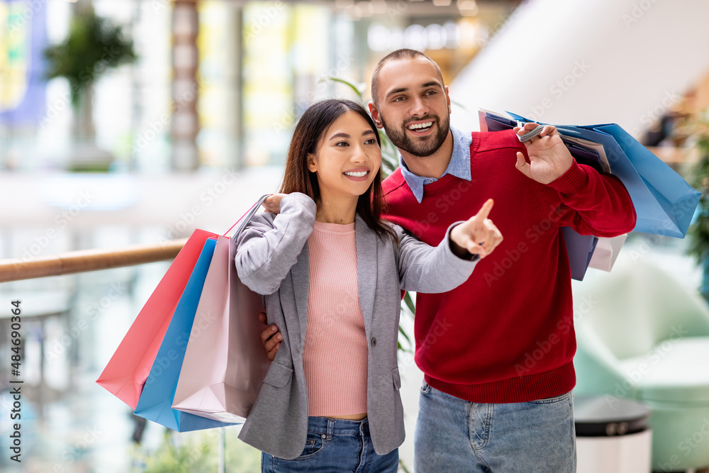 Shopping time. Young diverse couple holding bright paper bags, pointing at new shop at city mall Shopping time. Young diverse couple holding bright paper bags, pointing at new shop at city mall