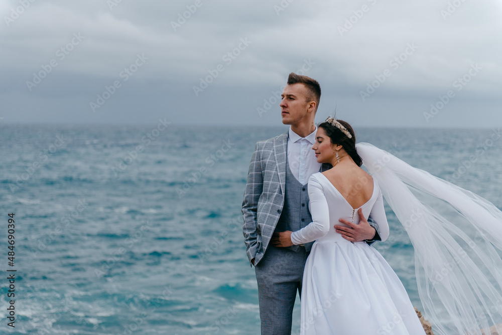 A young happy couple, a groom in suit, and a bride in white dress with a veil hugging and looking to one side. Blue sea background. Wedding shoot in Cyprus