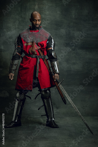 Vintage style portrait of brutal dark skinned man, medieval warrior or knight with wounded face wearing armour isolated over dark background. Comparison of eras, history