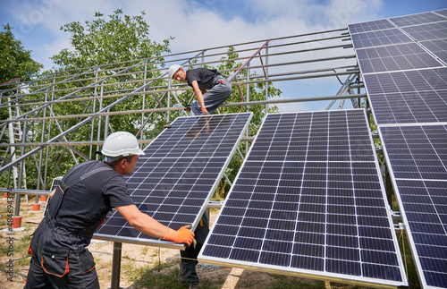 Male workers building photovoltaic solar panel system outdoors. Men engineers placing solar module on metal rails, wearing construction helmets and work gloves. Renewable and ecological energy.