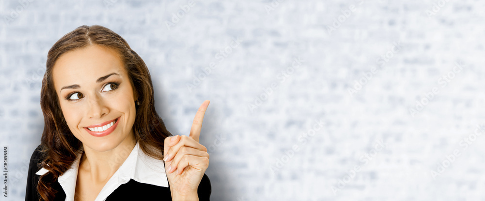 Smiling young business woman in black suit, pointing showing indicating copy space. Brunette businesswoman, on white bricks loft wall background Smiling young business woman in black suit, pointing showing indicating copy space. Brunette businesswoman, on white bricks loft wall background