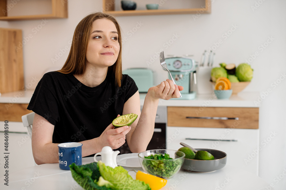 Young beautiful vegetarian girl dressed in pajamas eating fruits and vegetables for breakfast at home in the kitchen Young beautiful vegetarian girl dressed in pajamas eating fruits and vegetables for breakfast at home in the kitchen