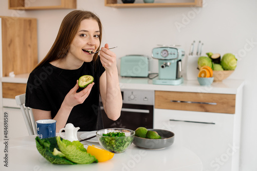 Young beautiful vegetarian girl dressed in pajamas eating fruits and vegetables for breakfast at home in the kitchen