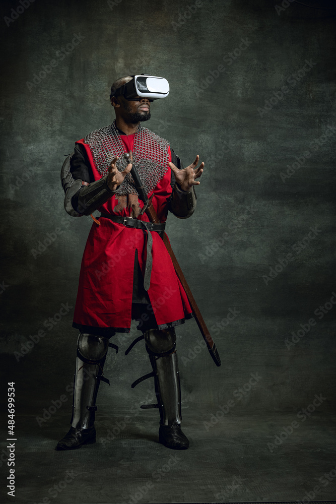 Naklejka premium Modern gadgets. One dark skinned man, medieval warrior or knight using VR headsets isolated over dark vintage background. Comparison of eras, history, technology