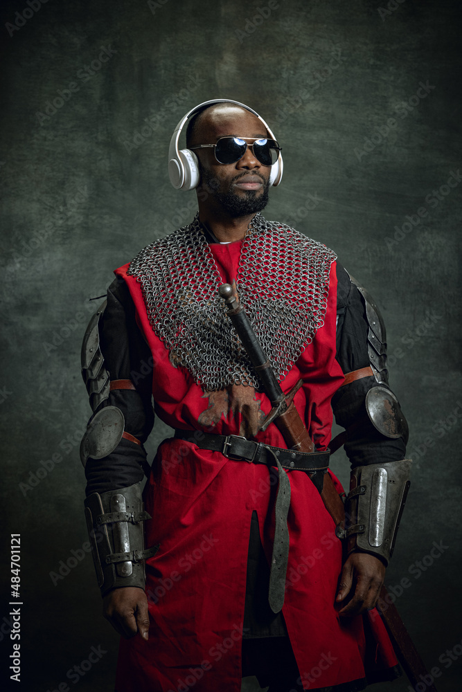 Vintage style portrait of stylish dark skinned man, medieval warrior or knight in sunglasses and headphones isolated over dark background. Comparison of eras, history