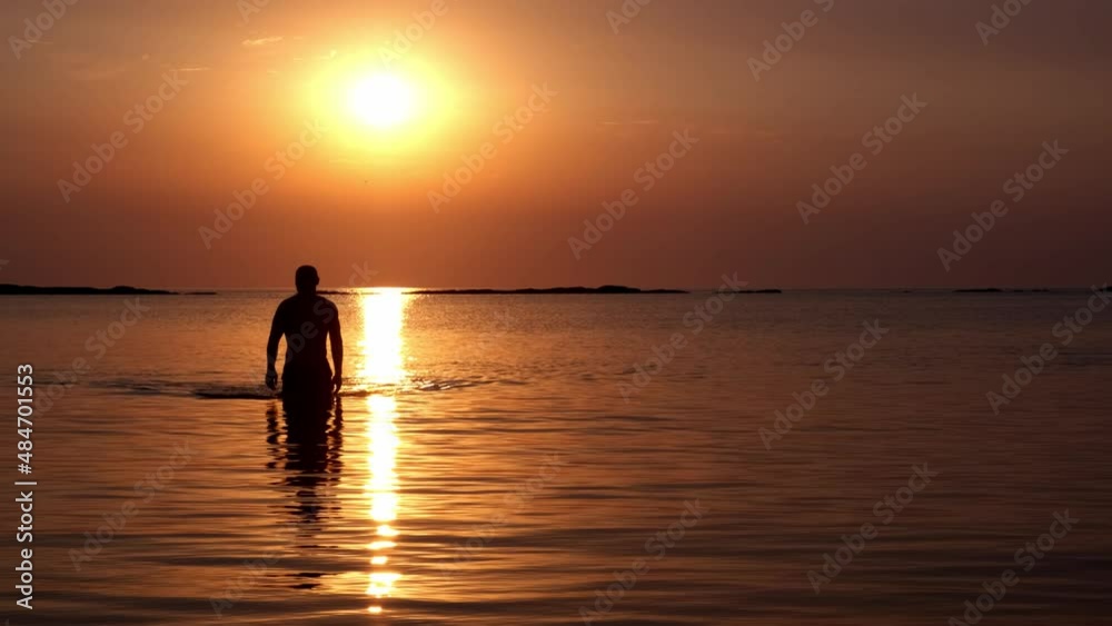 The silhouette of a man with an athletic body comes out of the sea water at sunset against the background of the sun disk in slow motion The silhouette of a man with an athletic body comes out of the sea water at sunset against the background of the sun disk in slow motion