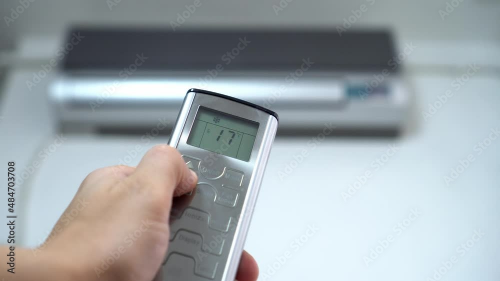 A man's hand with a remote control is aimed at the air conditioner. By moving your finger and pressing the button, the numbers change on the display, cooling and lowering the temperature. hot. A man's hand with a remote control is aimed at the air conditioner. By moving your finger and pressing the button, the numbers change on the display, cooling and lowering the temperature. hot.