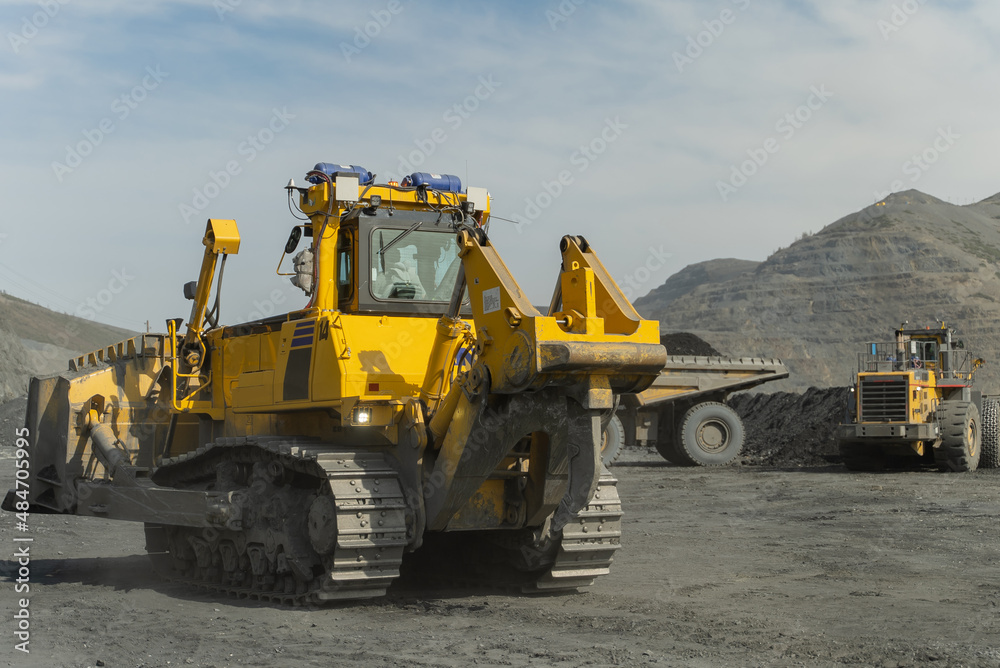 A bulldozer drives through an open quarry. The ore is being loaded in the background. A bulldozer drives through an open quarry. The ore is being loaded in the background.