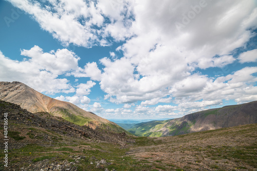 Scenic view from mountain pass to green forest valley among mountain ranges and hills on horizon at changeable weather. Green landscape with sunlit mountain vastness under cumulus clouds in blue sky.