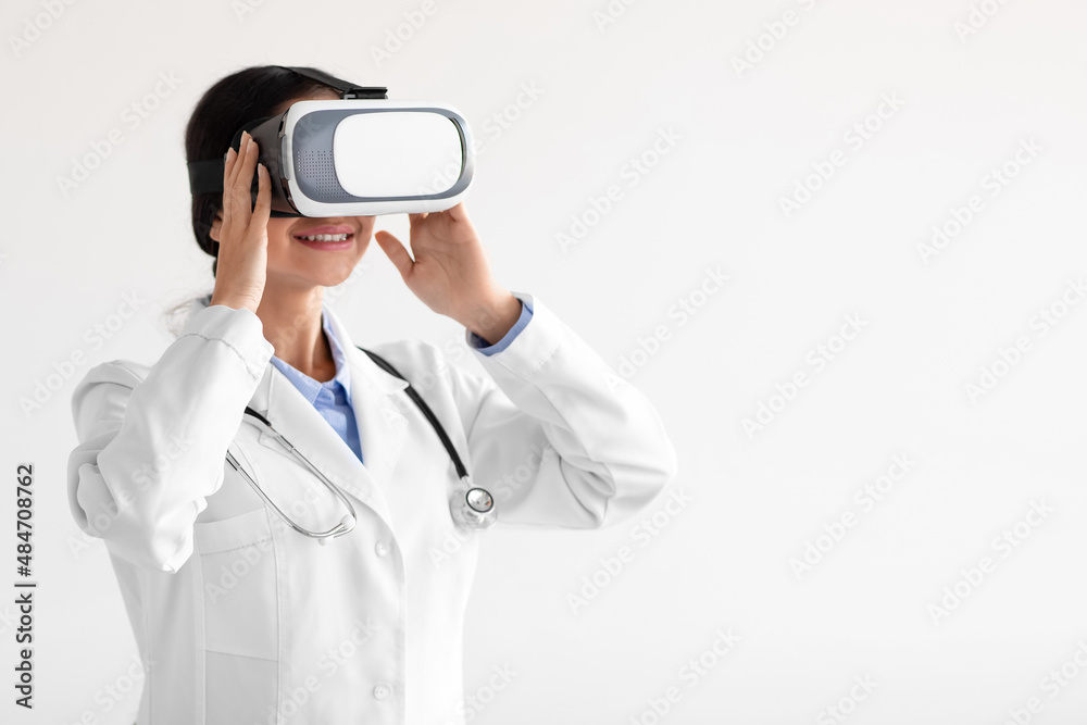 Smiling millennial indian female doctor in white coat and virtual reality glasses isolated on light background