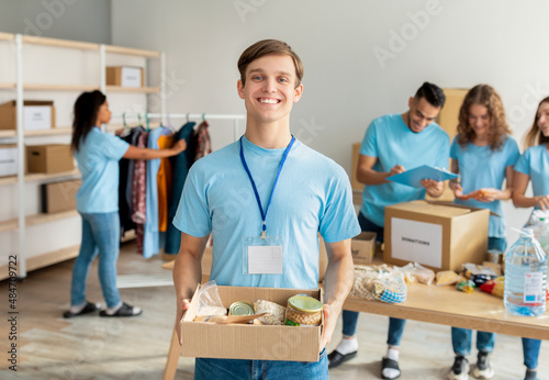 Happy male activist volunteer in blue uniform holding cardboard box with food fo Fototapet