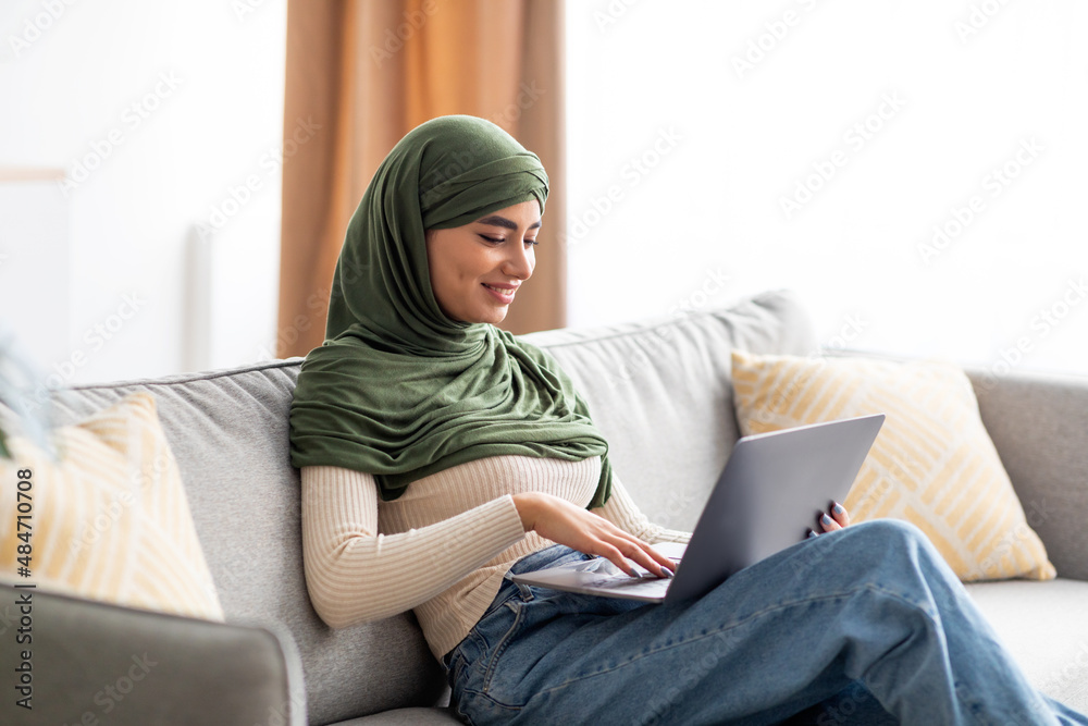 Portrait of young Arab female in hijab sitting on sofa, using laptop, surfing web, working or studying remotely at home Portrait of young Arab female in hijab sitting on sofa, using laptop, surfing web, working or studying remotely at home