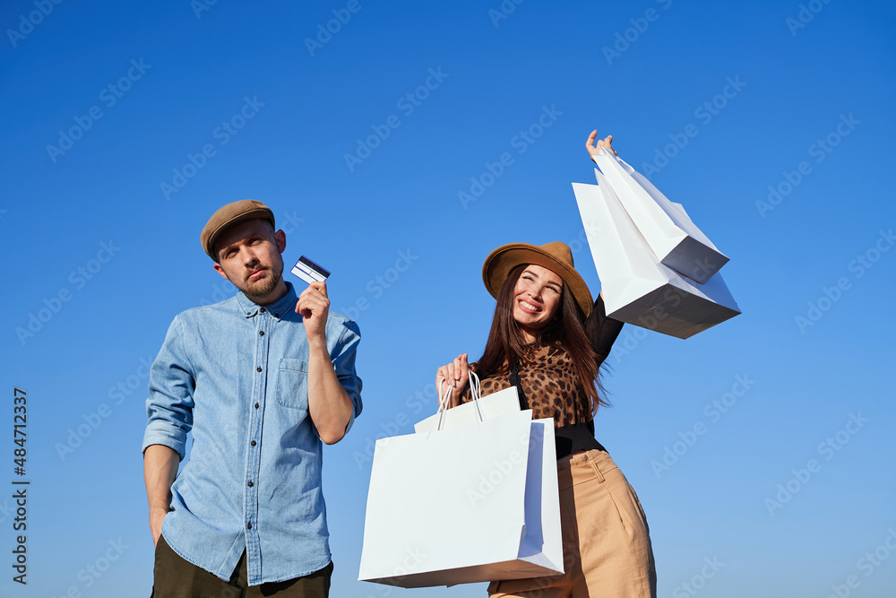 Sales, discounts or black friday concept. Outdoor portrait of attractive fashion styled couple: male with credit card and woman with shopping bags in hands posing together over blue sky background