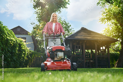 Fotografie Low angle view of smiling caucasian woman with blond hair using electric lawn mower while working at garden