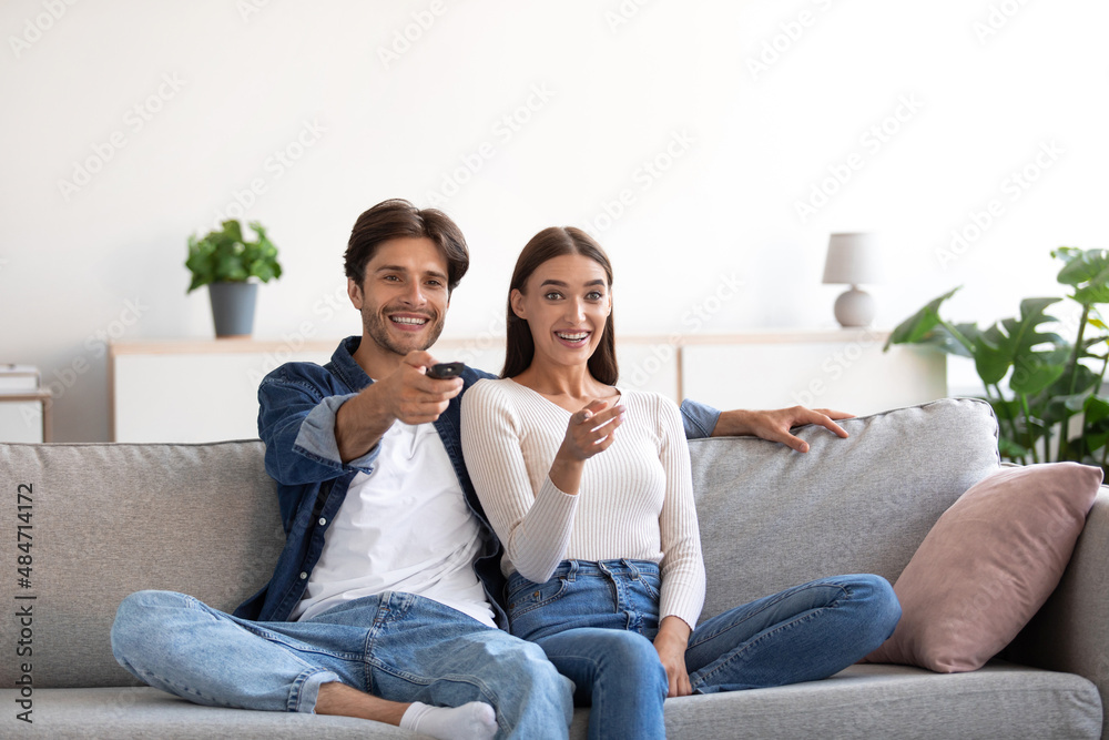 Happy laughing relaxed millennial european woman and man with remote control hugging, watching tv