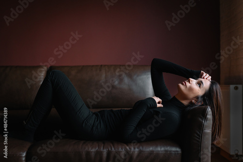 Middle age pensive woman with dark hair in black clothes lie on brown leather sofa putting hand on forehead, looking at ceiling. Depression, fatigue, stress, problems, emotions concept. Vertical. 