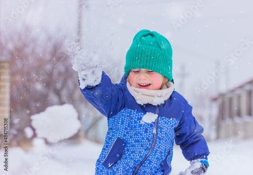 cute child boy in a knitted green hat and white scarf on a snowy street playing snowballs. winter holidays, outdoor activities in the snow, childhood.