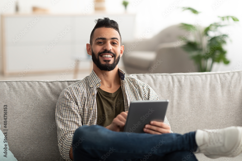 Handsome young Arab man using digital tablet while sitting on couch at home Handsome young Arab man using digital tablet while sitting on couch at home