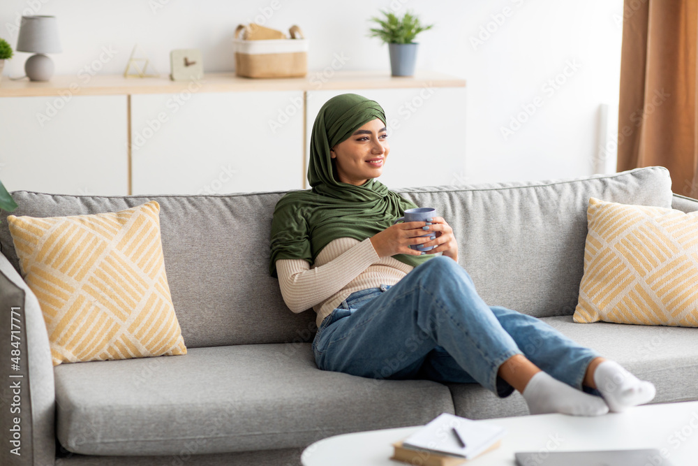 Obraz premium Pretty young Arab lady in hijab sitting on sofa with hot drink, spending peaceful weekend at home, copy space