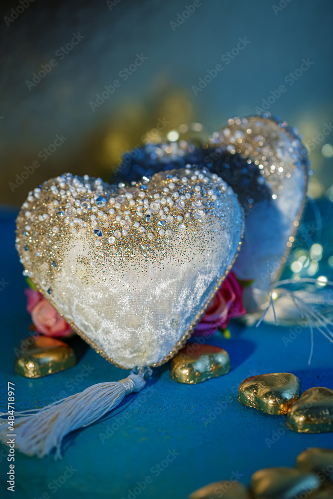 Two velvet white hearts with beads and golden foil chocolates on a blue table. Dark blurred background with bokeh. Flowers. Two velvet white hearts with beads and golden foil chocolates on a blue table. Dark blurred background with bokeh. Flowers.