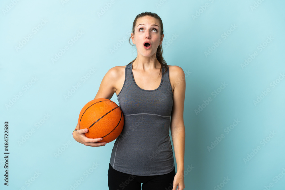 Young woman playing basketball isolated on blue background looking up and with surprised expression Young woman playing basketball isolated on blue background looking up and with surprised expression