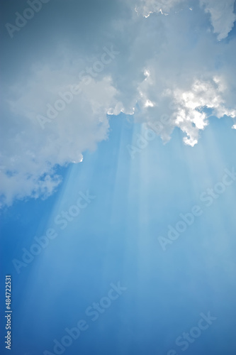 Sun rays beaming through white clouds against a bright blue sky. 