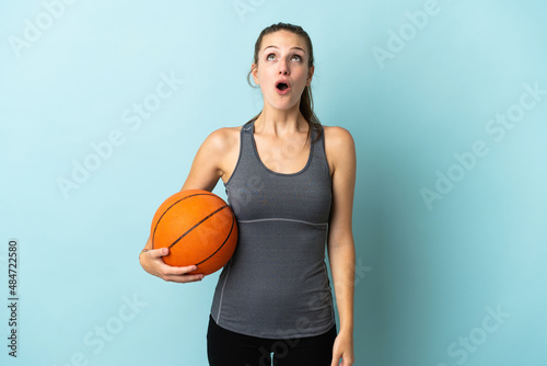 Young woman playing basketball isolated on blue background looking up and with surprised expression