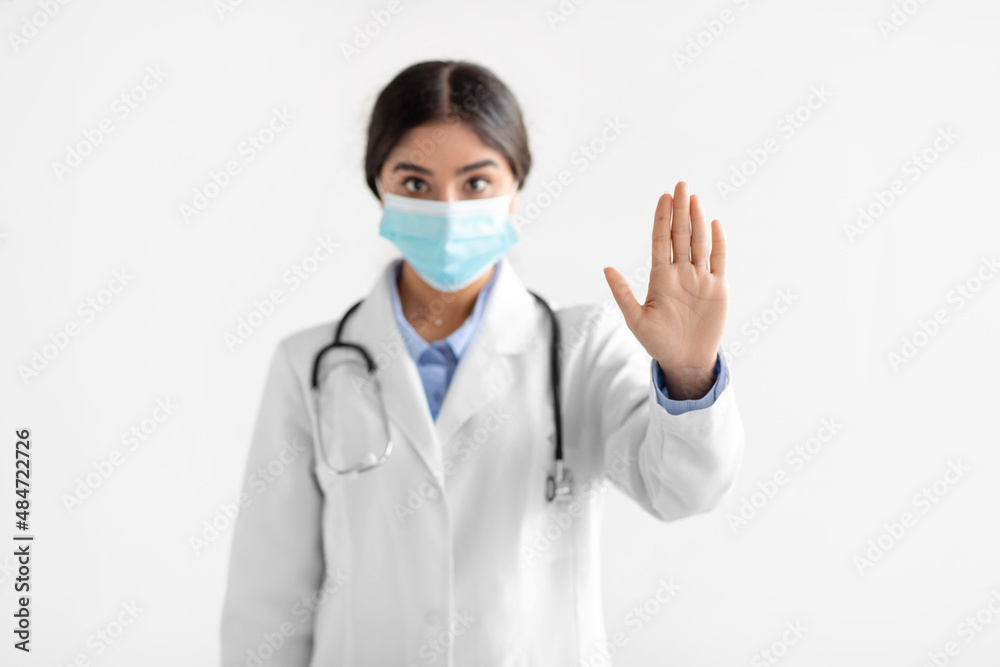 Serious young indian woman doctor in coat and protective mask show stop hand gesture