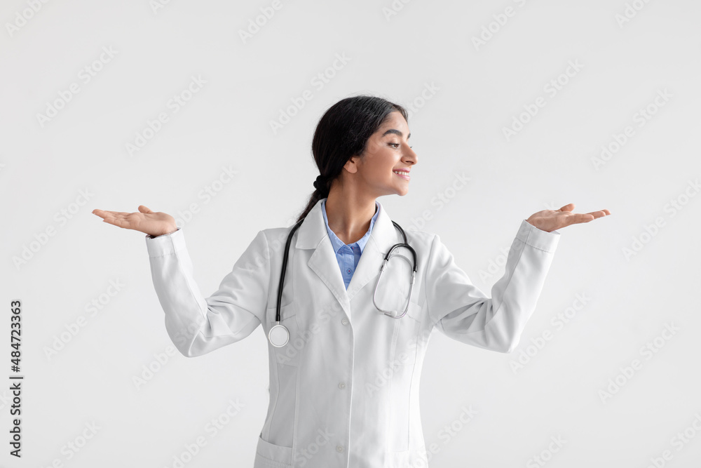Smiling millennial indian woman doctor in white coat raises her hands and makes choice, isolated on light background Smiling millennial indian woman doctor in white coat raises her hands and makes choice, isolated on light background