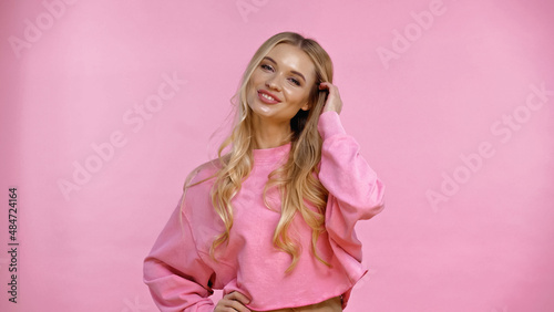Happy blonde woman touching hair isolated on pink.