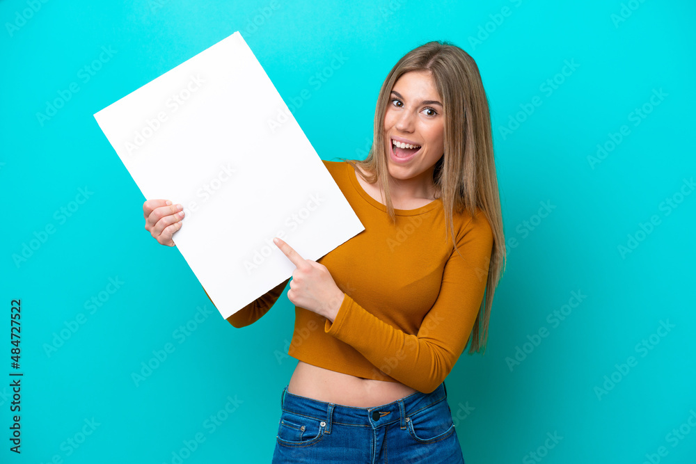 Young caucasian woman isolated on blue background holding an empty placard with happy expression and pointing it Young caucasian woman isolated on blue background holding an empty placard with happy expression and pointing it