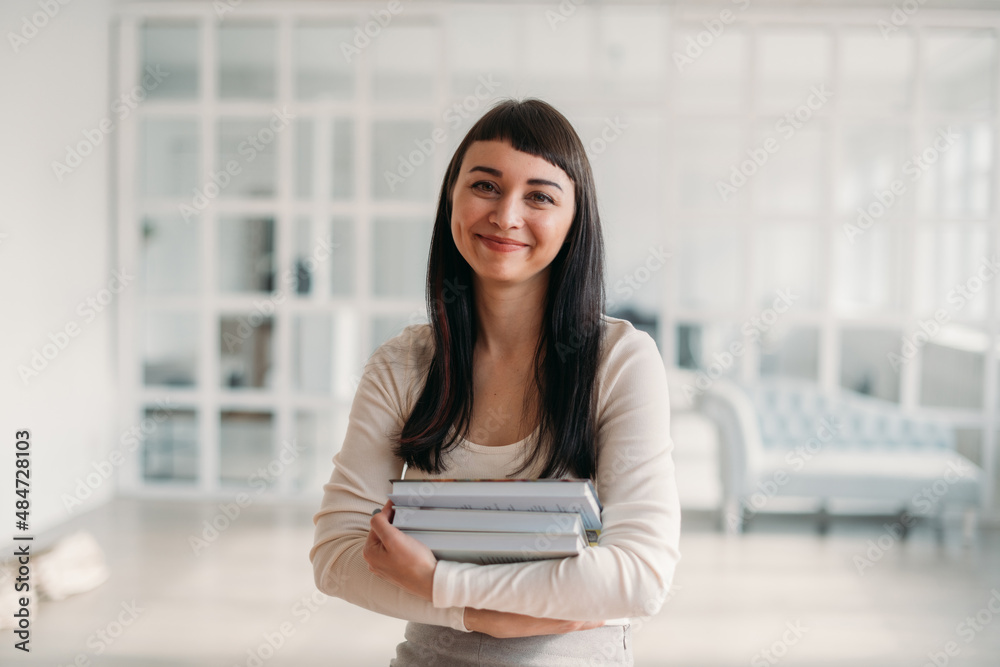 Portrait of a young beautiful woman with books in her hands in a large bright room. Beautiful brunette, young teacher or student