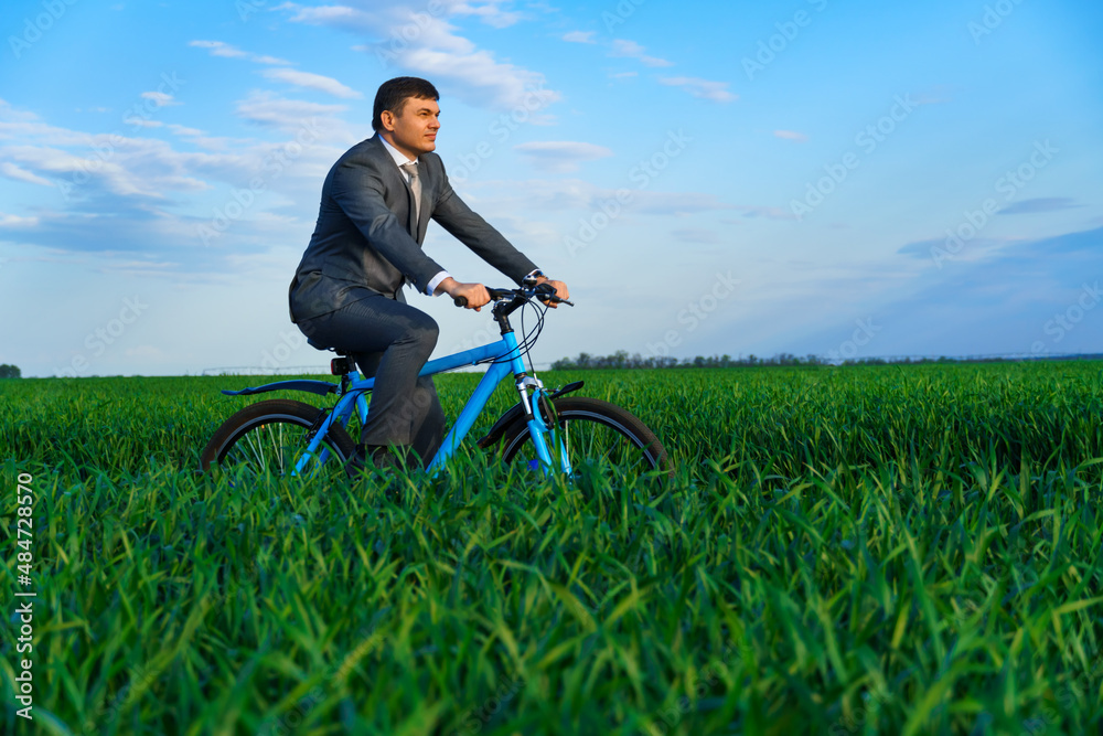 business concept - A businessman rides a bicycle on a green grass field, dressed in a business suit, he has a briefcase and documents business concept - A businessman rides a bicycle on a green grass field, dressed in a business suit, he has a briefcase and documents