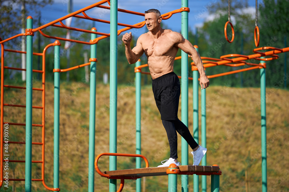 Active shirtless man doing exercises for legs on bench on sports ground. Caucasian bodybuilder with muscular torso training regularly for keeping sporty body shape. 