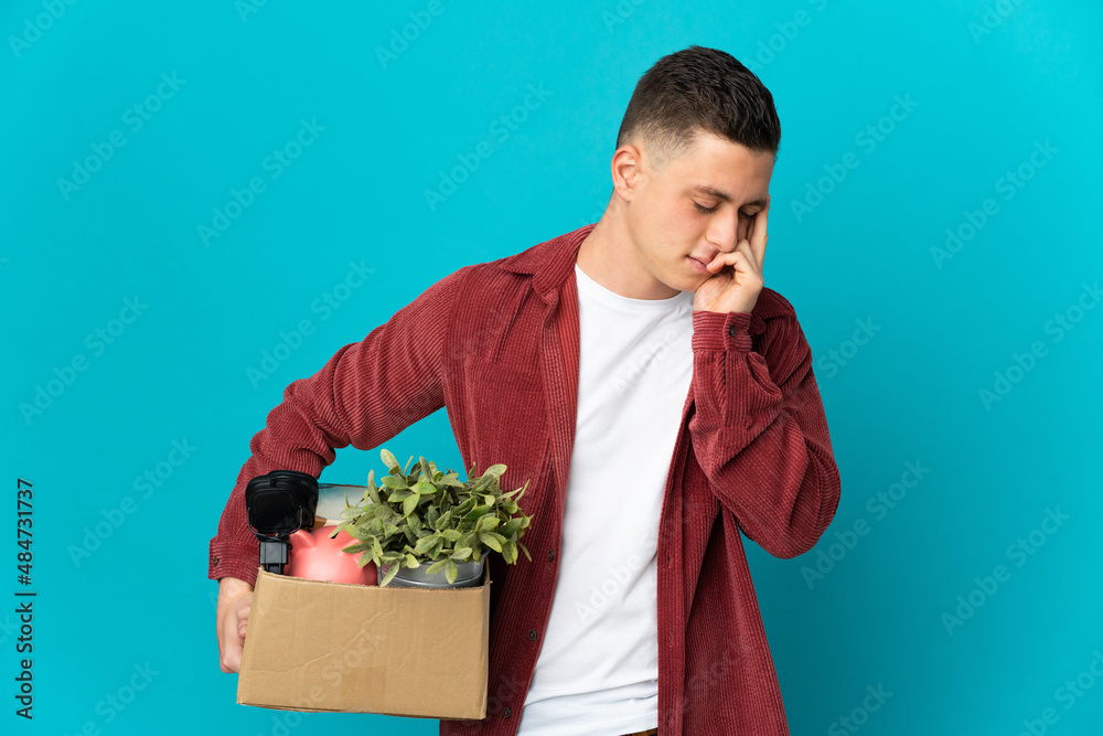 Young caucasian man making a move while picking up a box full of things isolated on blue background with headache Young caucasian man making a move while picking up a box full of things isolated on blue background with headache